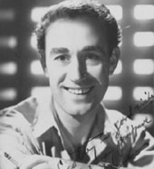 Black and white portrait of a smiling man with short curly hair, wearing a light-colored shirt, in front of a backdrop with horizontal light patterns.